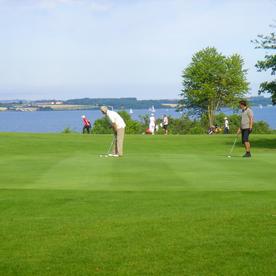 Zwei Golfer im Vordergrund auf dem Green, im Hintergrund die Flensburger Förde.