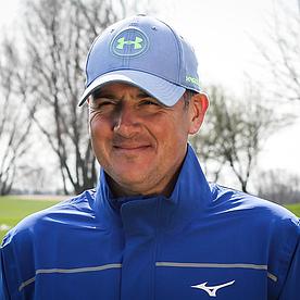 Ein Golftrainer mit blauem Anzug und blauer Cap vor einem Green.